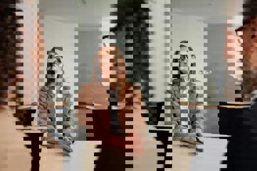 Kvinna med glasögon i en brun kostym vid ett bord framför två personer.